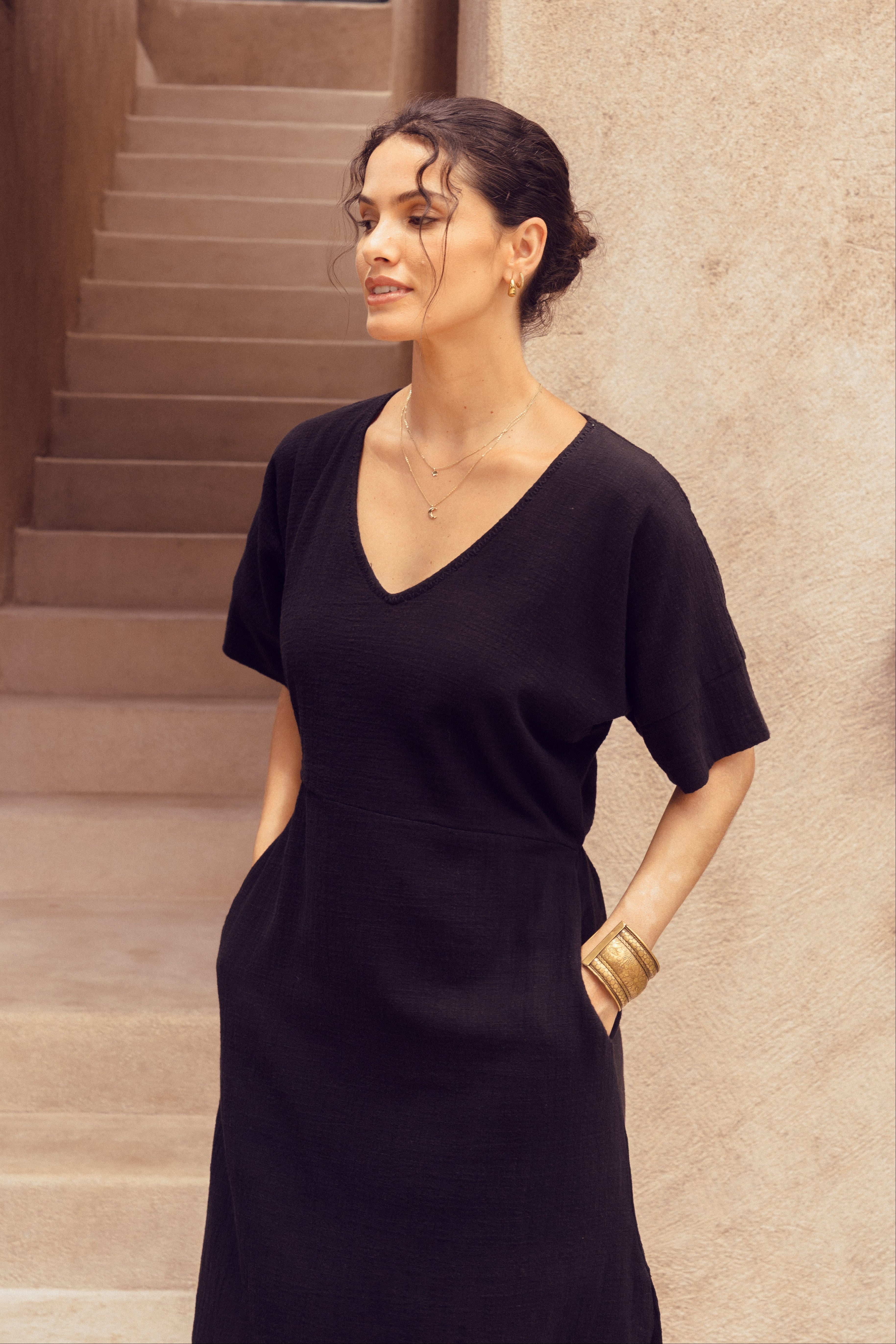 Woman in a black dress standing in a sunlit hallway with beige walls and steps.
