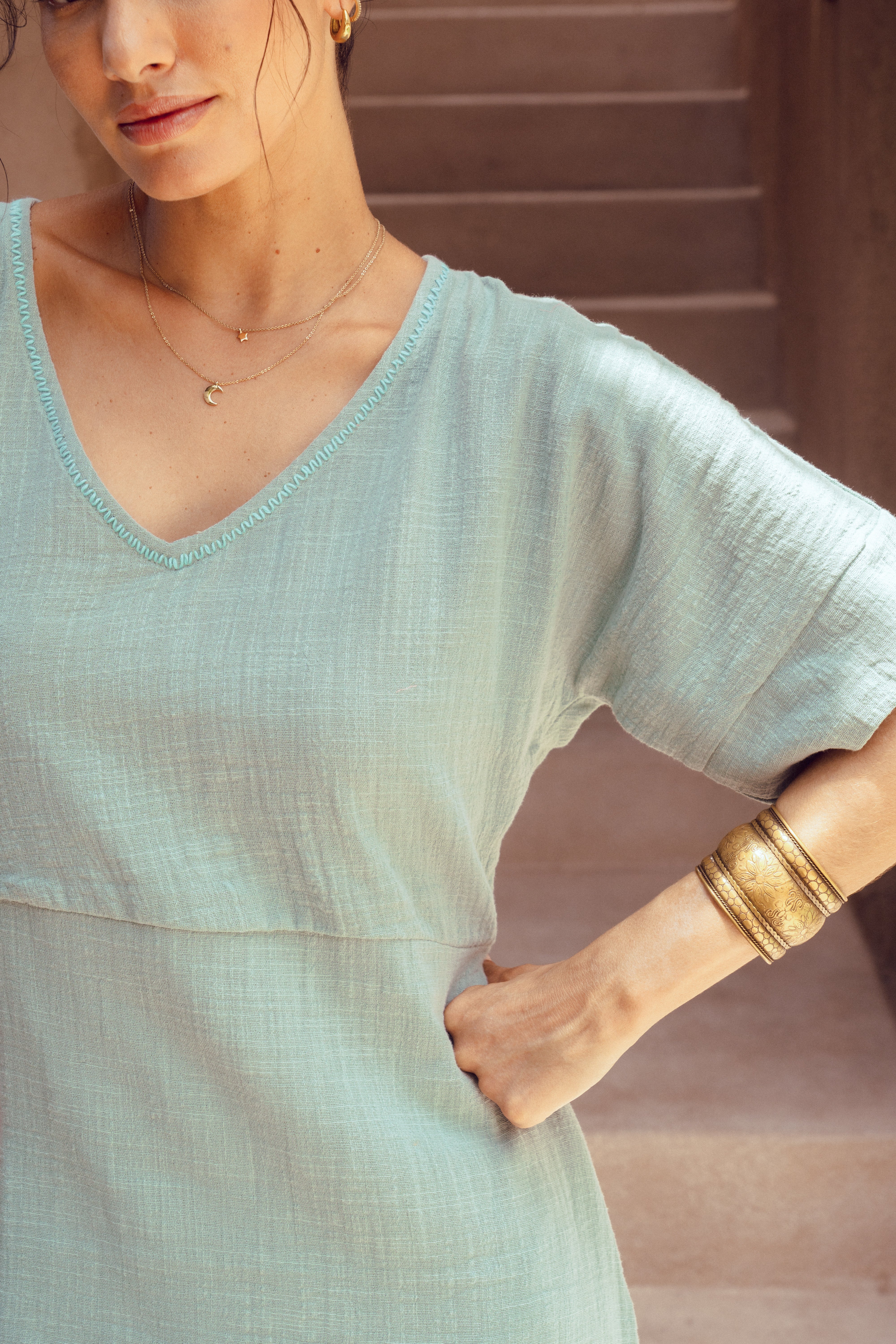 Woman wearing a light blue dress with gold jewelry against a neutral background