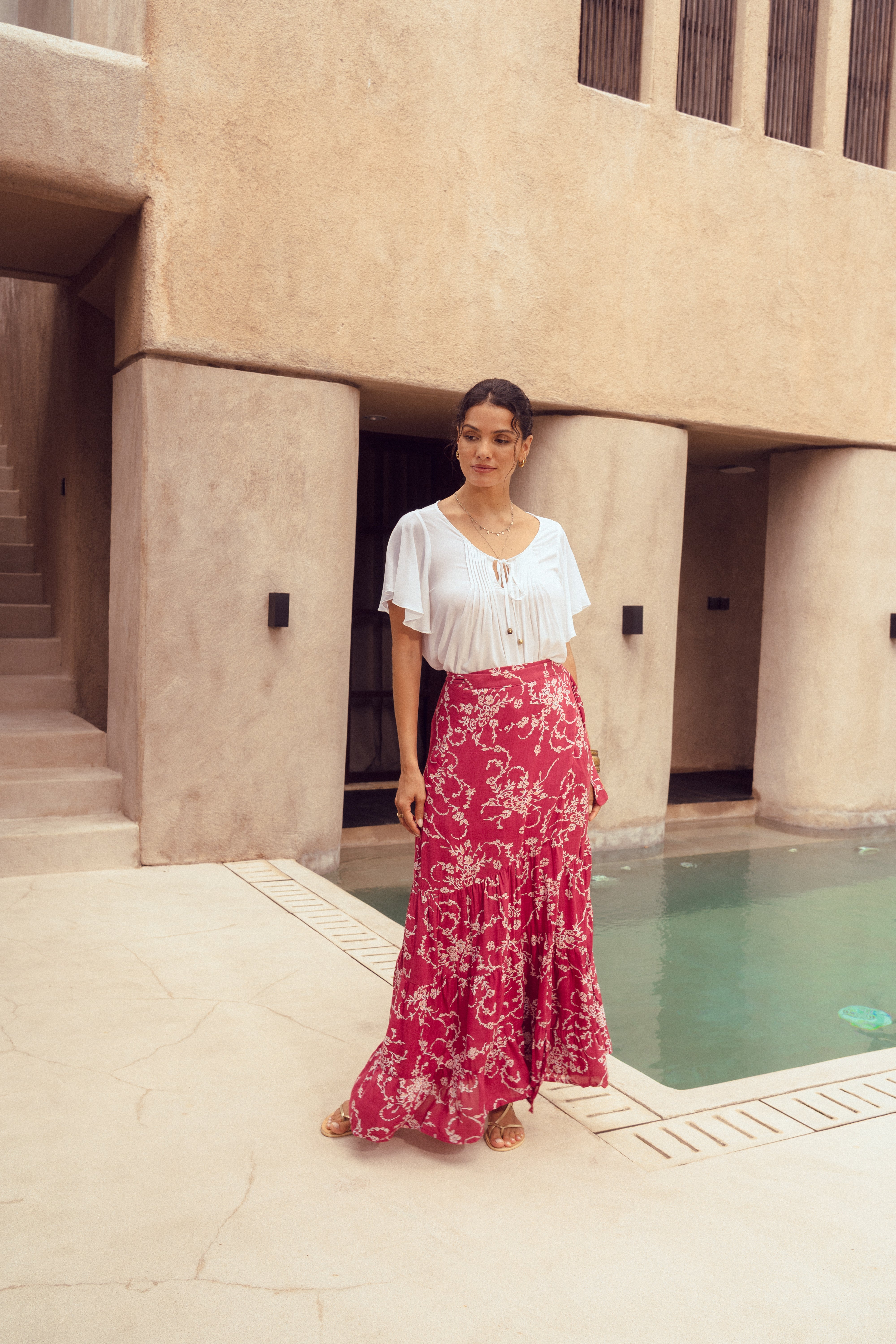 Woman in a white blouse and red floral skirt standing by a poolside.