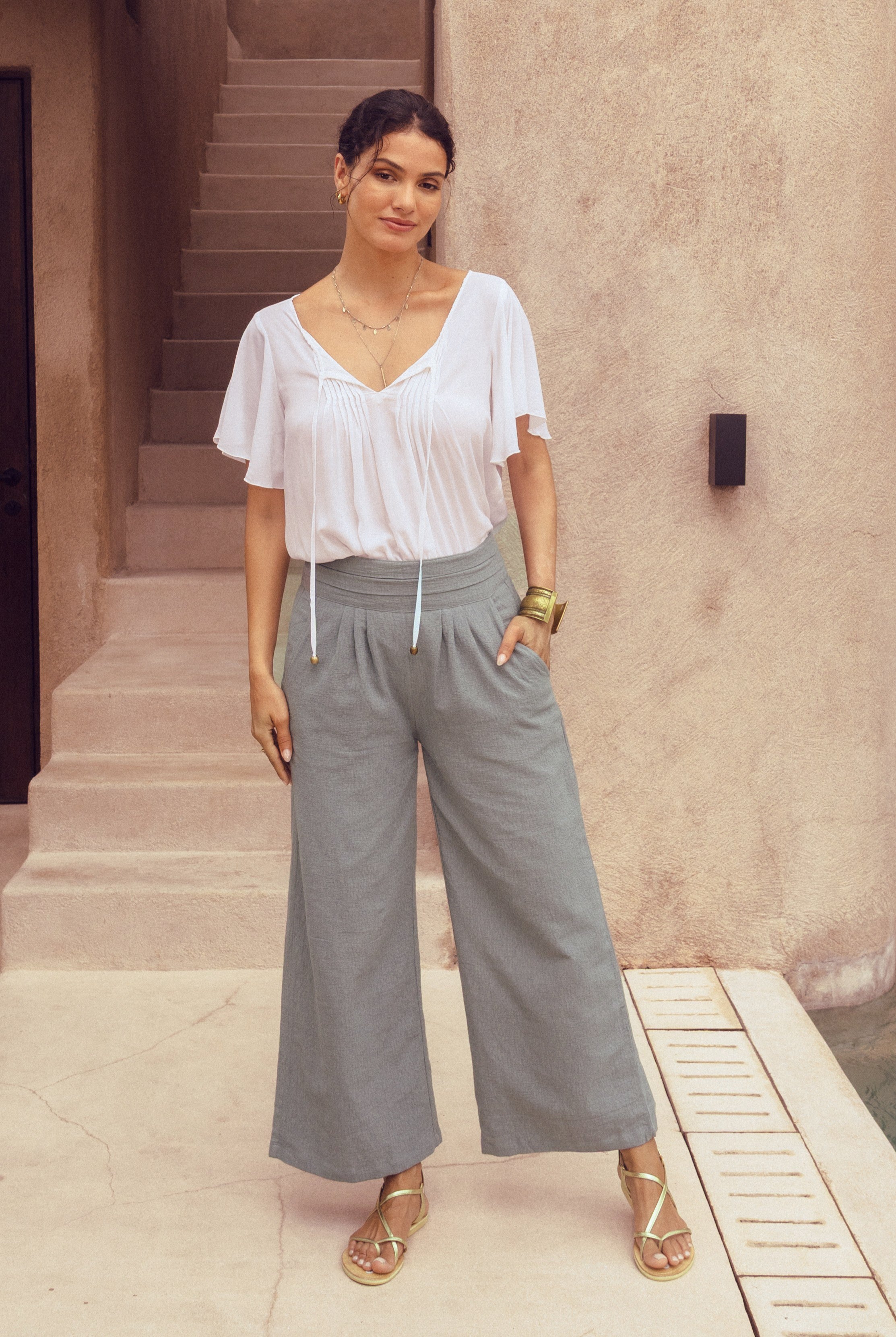 Woman wearing a white blouse and gray wide-leg pants standing in front of a staircase.