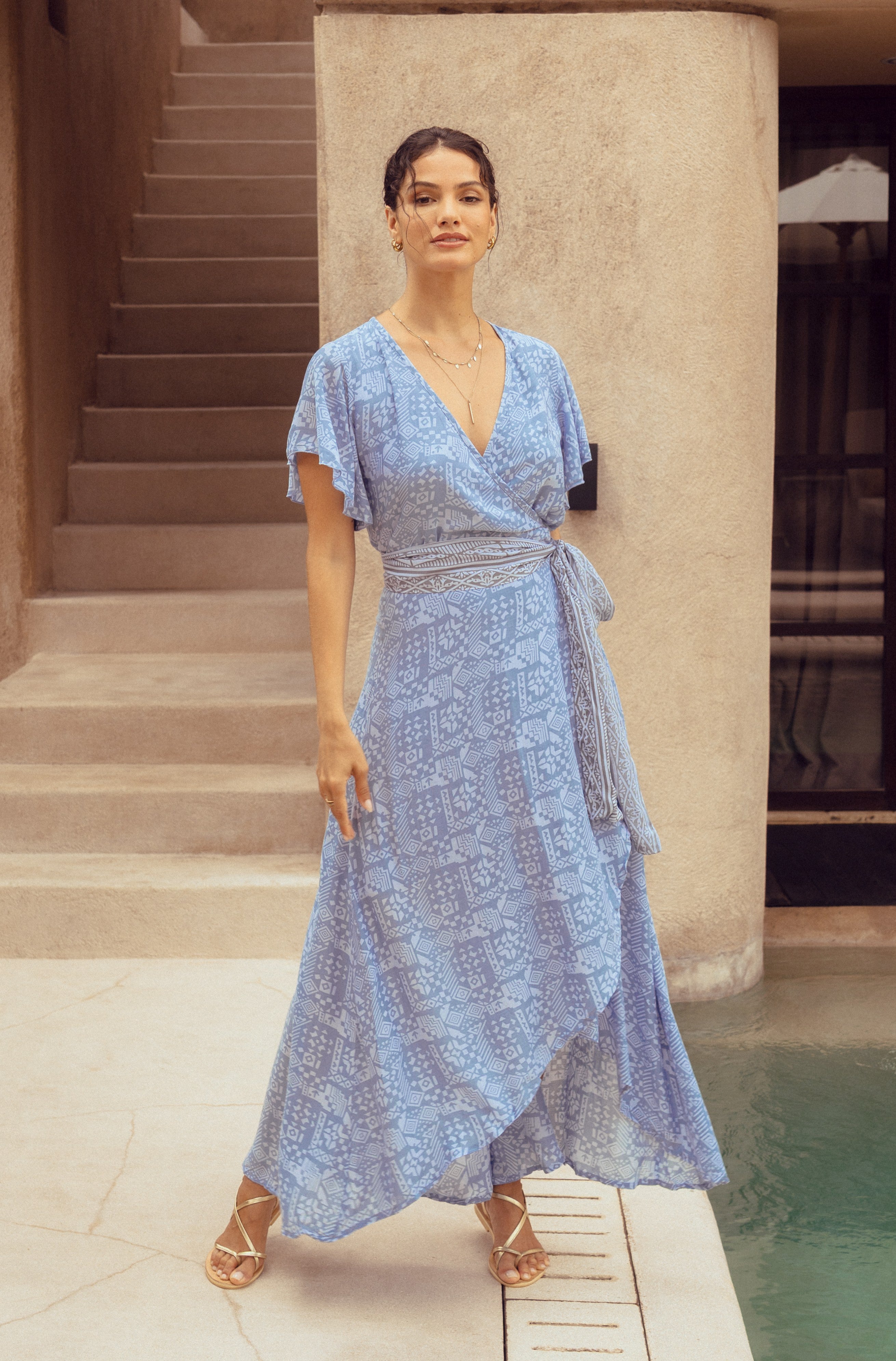 Woman in a blue patterned wrap dress standing by a poolside.