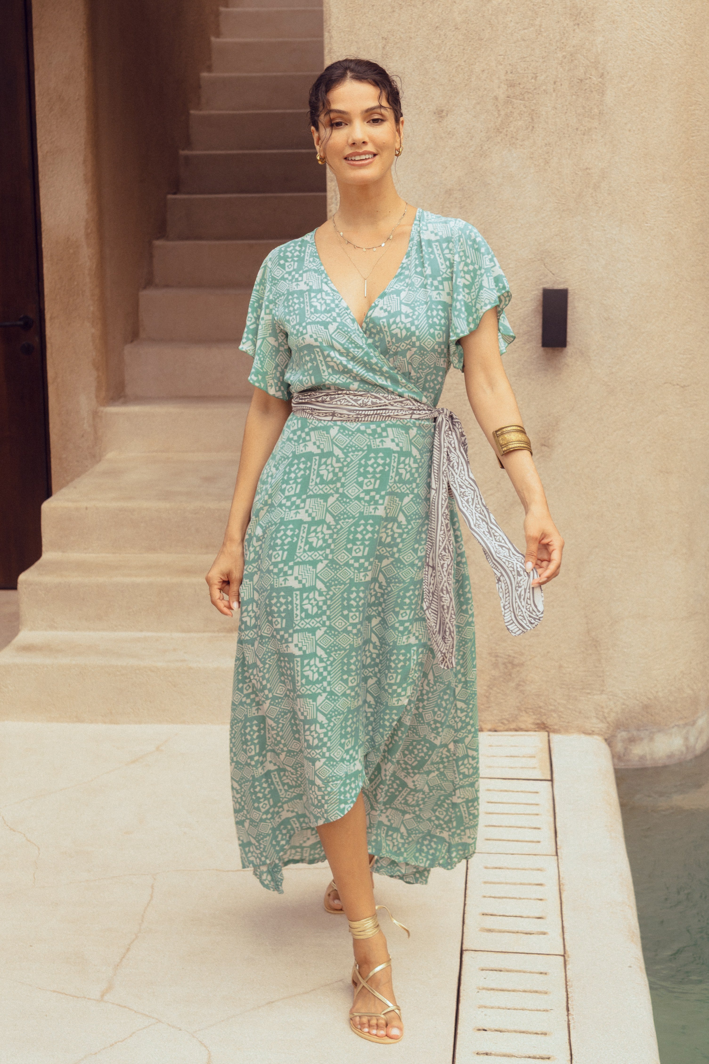 Woman in a green patterned wrap dress standing on steps outdoors.