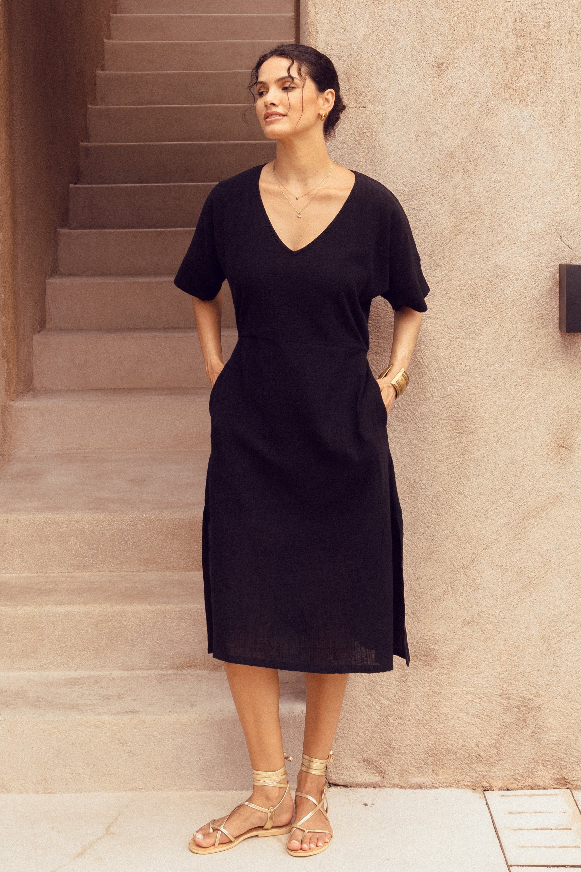 Woman in a black cotton dress standing on a staircase in front of a beige building.