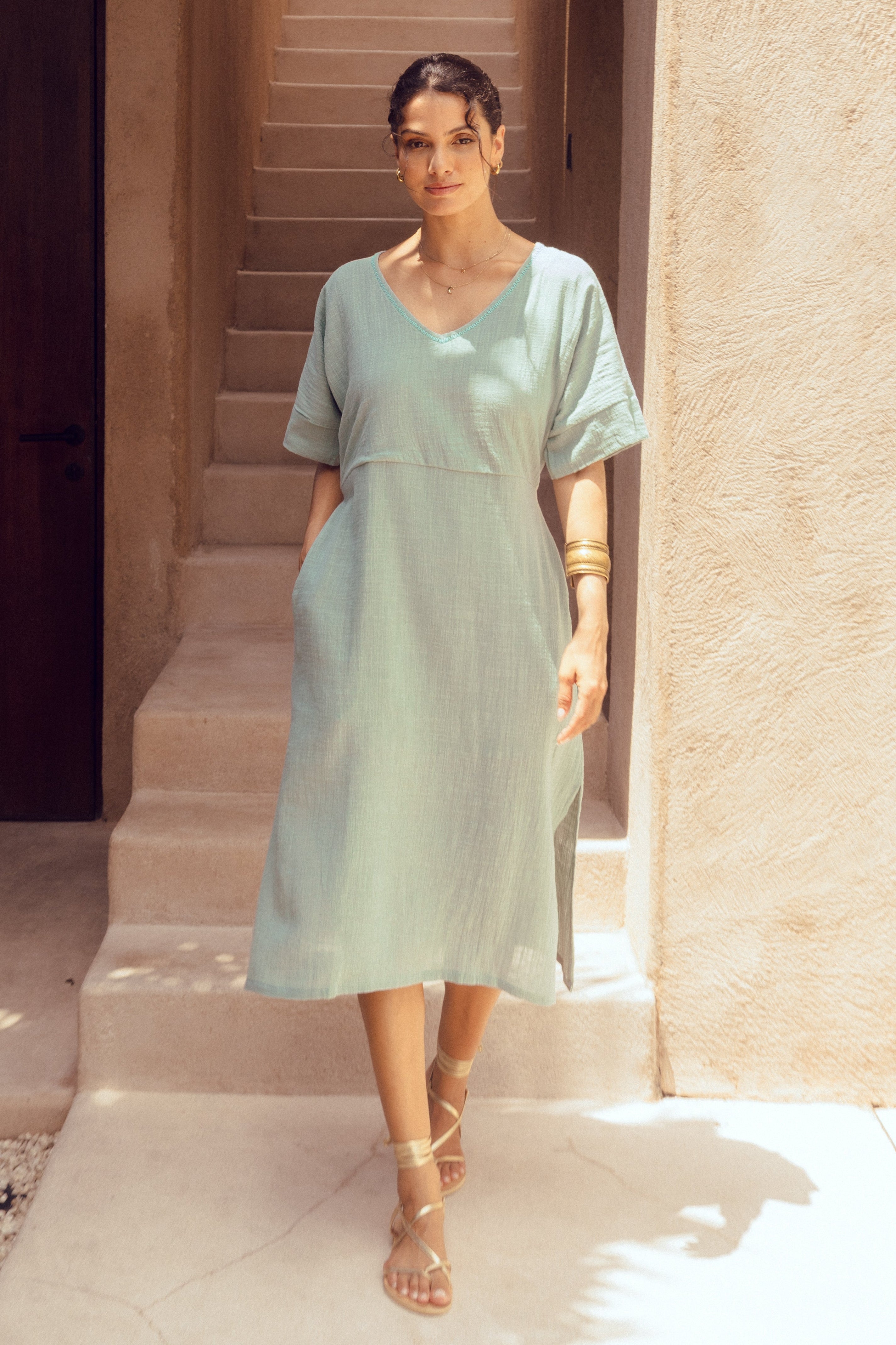 Woman in a light blue dress standing in a sunlit outdoor setting with stone walls.