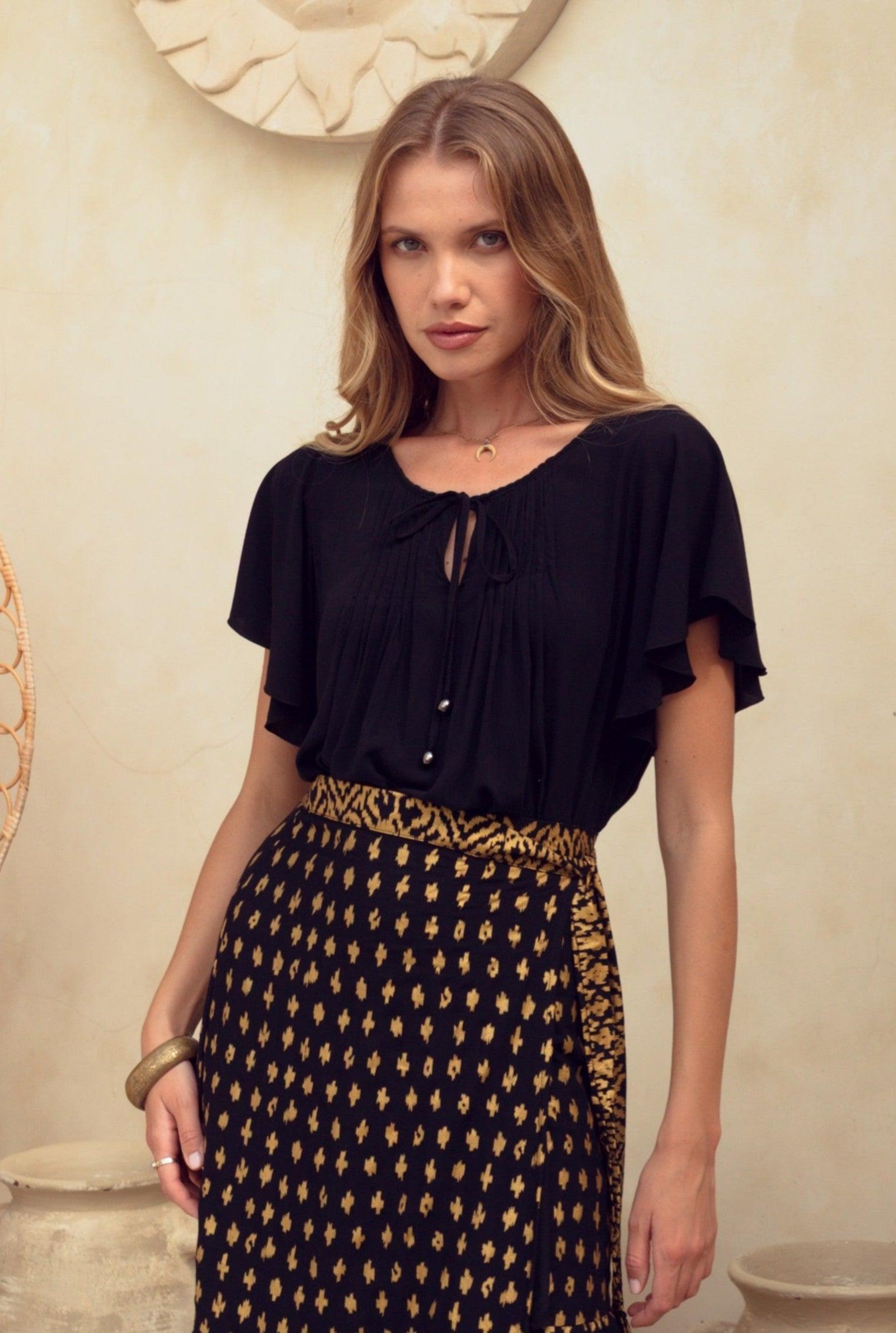 Woman wearing a black top and patterned skirt standing in a decorative room with vases and a chair.