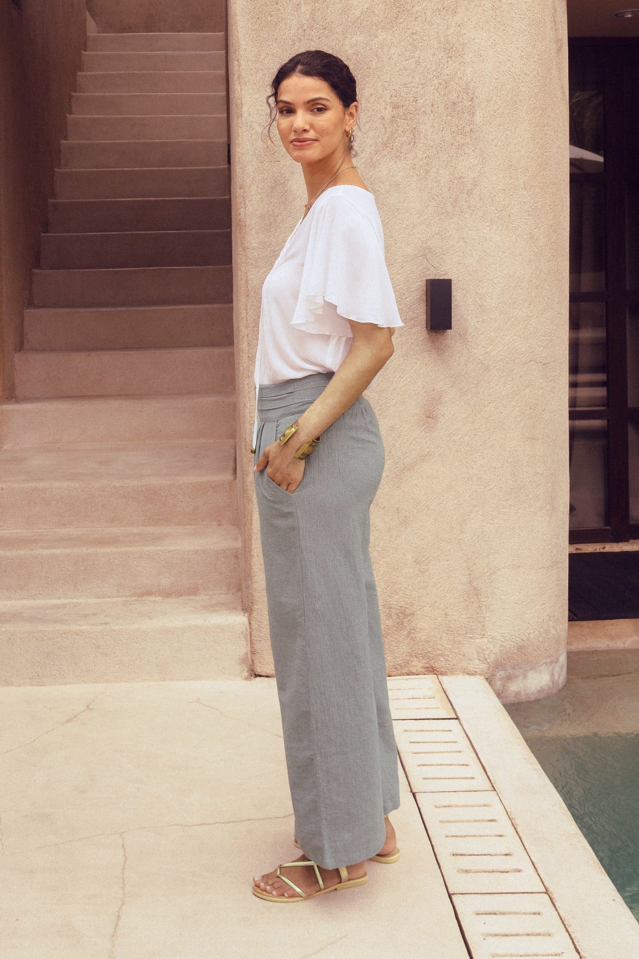 Woman in a white top and gray pants standing by a building with steps and a pool.