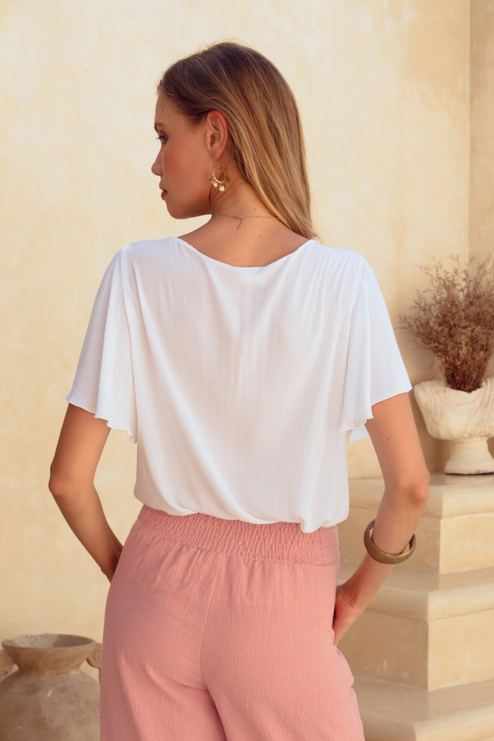 Woman in a white top and pink pants standing in a sunlit outdoor setting with a cactus plant and stone steps.