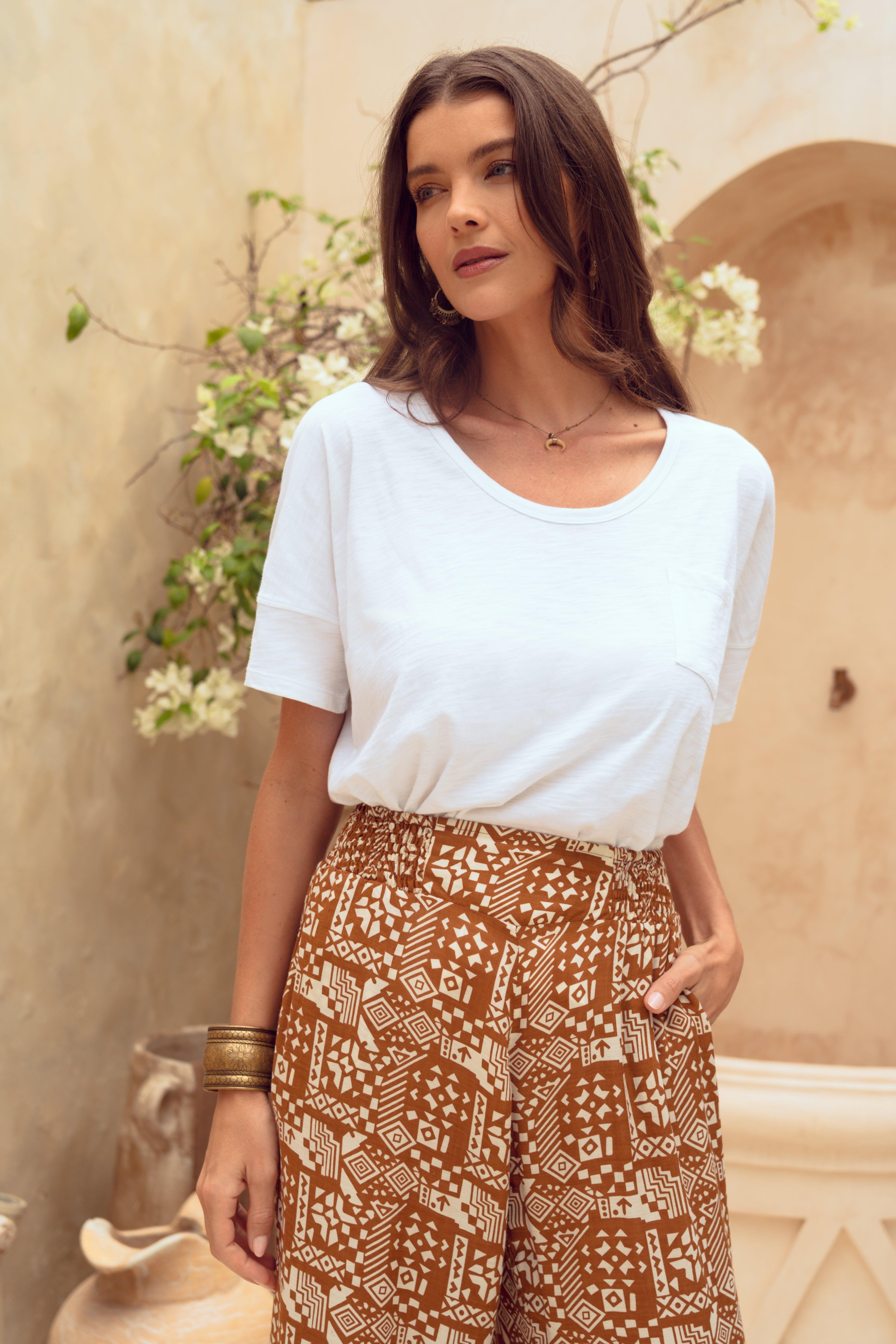 Woman wearing a white top and patterned brown pants standing against a beige wall with greenery.