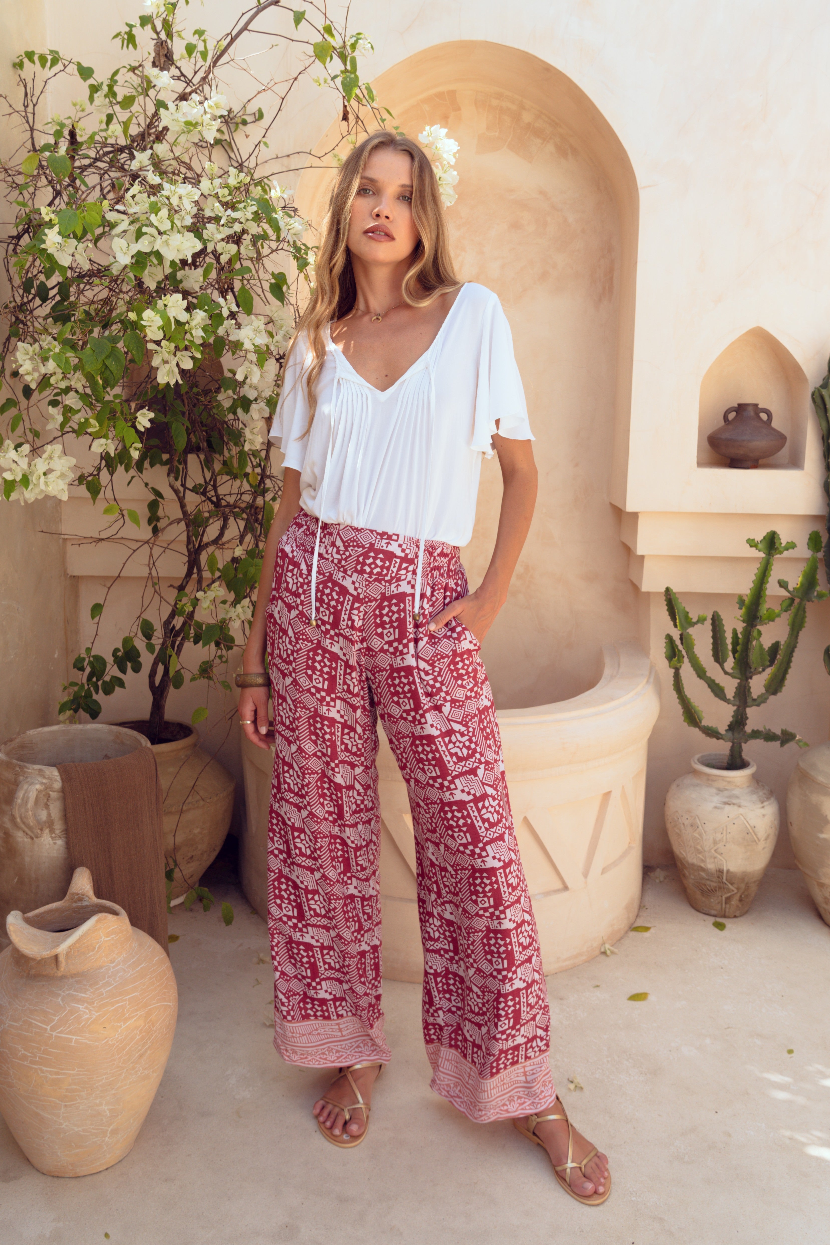 Woman wearing a white top and red patterned pants standing in a decorative outdoor setting with plants and vases.