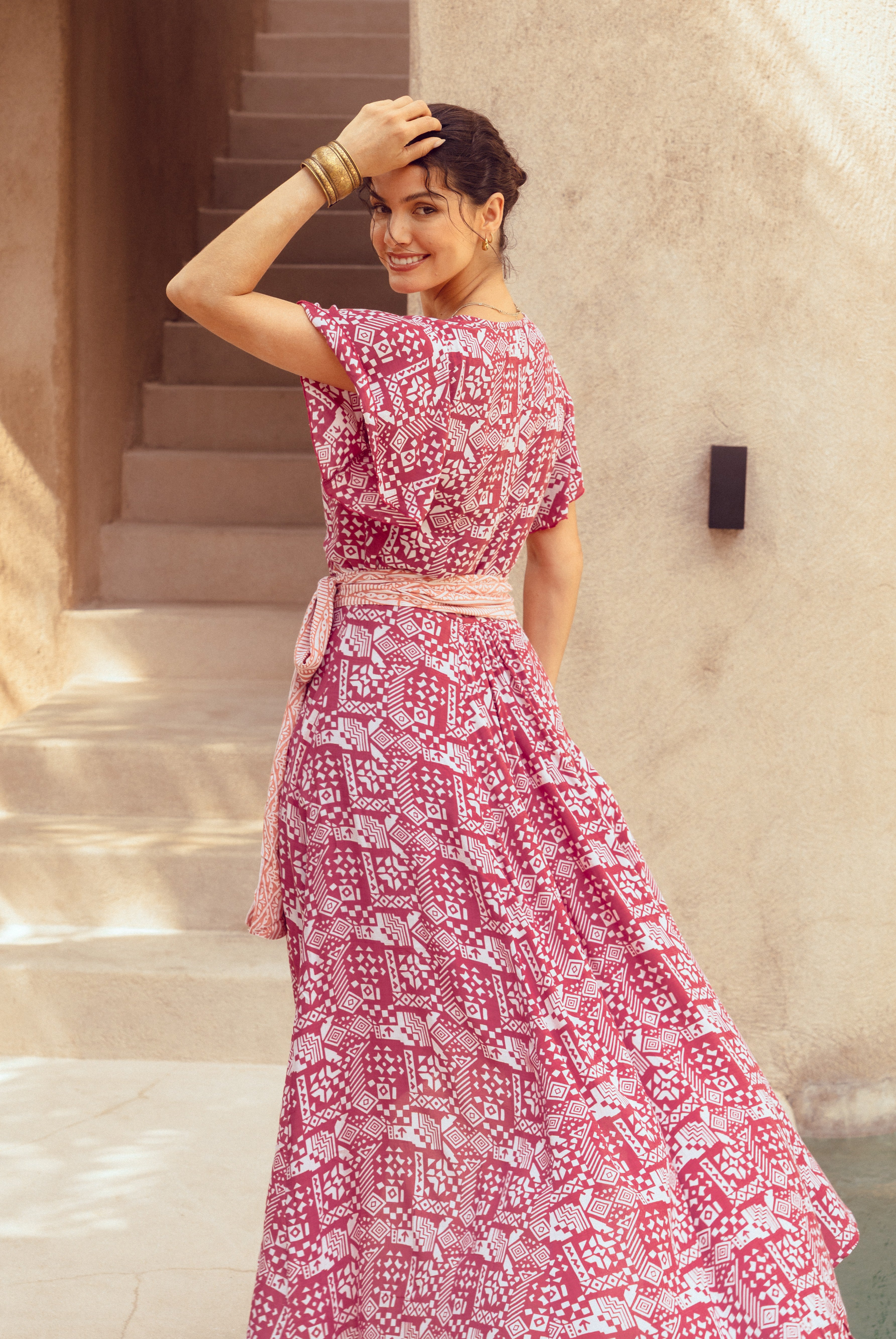 Woman in a pink patterned dress standing outdoors against a beige wall.