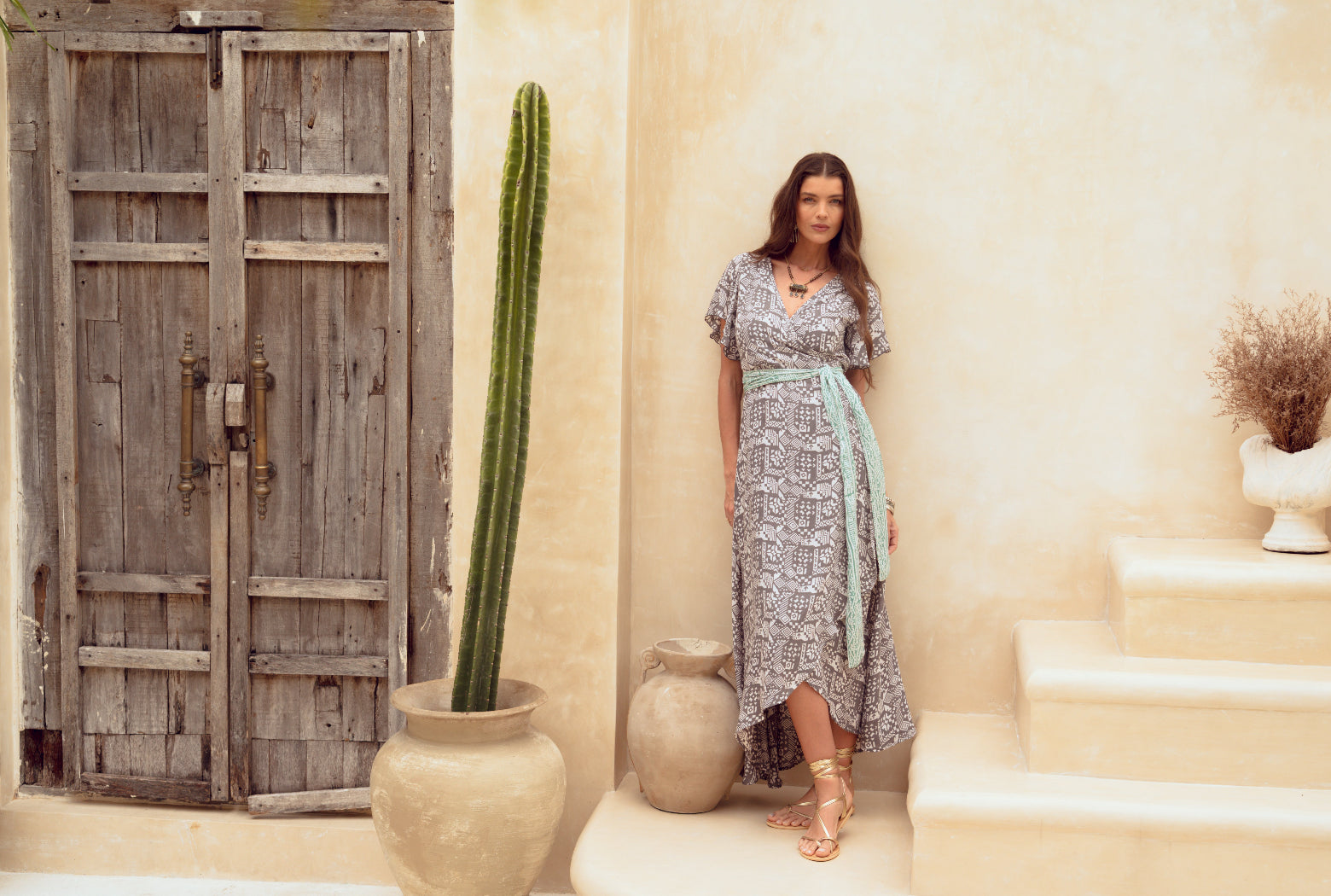 Woman in a patterned dress standing in a sunlit room with a cactus and decorative pots.